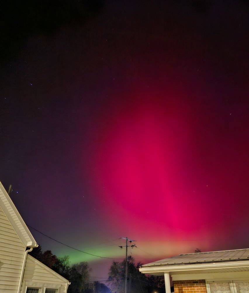 A photograph of the night sky over two houses, one brick and one with white siding. The sky is lit by a rosy red aurora with a ray running through it. A band of ghostly green aurora runs just above the horizon.