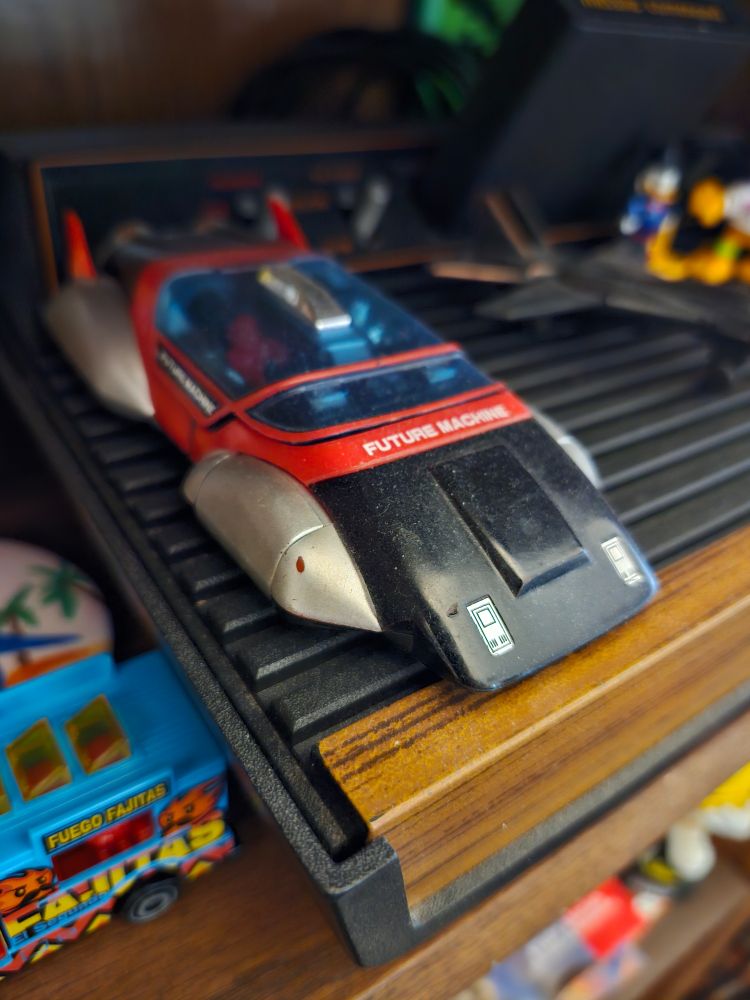 A color photograph featuring a close-up shot of Psycho, a GO BOTS toy. Psycho is shown here in car form, a streamlined, blue-tinted glass-roofed, futuristic-looking red, black, and silver vehicle labelled "FUTURE MACHINE." Psycho is parked on top of a woodgrained ATARI 2600. Other items visible in the photograph, in varying degrees of focus or blur, are a brass fighter jet pencil sharpener, a pair of figurines (Garfield and Scrooge McDuck), a blue Hot Wheels food truck labelled "Fuego Fajitas," and a pin with palm trees on it.