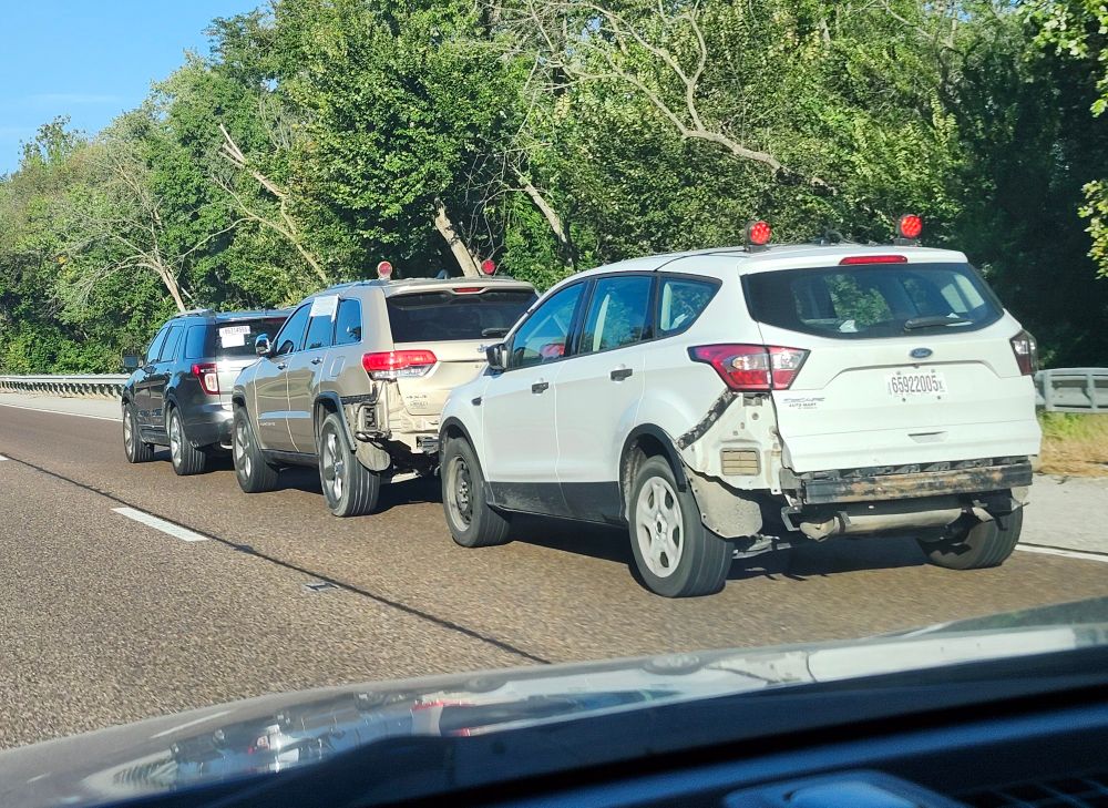 A photograph of a trio of vehicles travelling down the interstate. The first car is towing the two behind it, and the three are hitched together bumper-to-bumper in a fashion reminiscent of the victims in the first Human Centipede film, in which three people are stitched together, mouth-to-anus, to create 