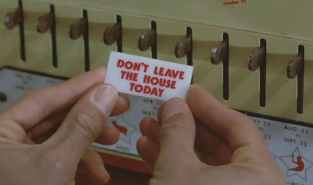A screenshot from the 1985 film Pee-Wee's Big Adventure. In extreme close-up, a pair of pale hands hold a small fortune dispensed from a vending machine  (in the background) bedecked with a series of knobs above calendar dates. The held fortune reads: "DON’T LEAVE THE HOUSE TODAY."