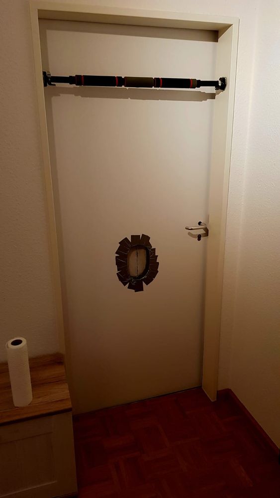 Mein Gloryhole in der Wohnung. Eine weiße Tür mit einem großen Gloryhole, die Ränder des Lochs mit Panzertape abgeklebt. Oben im Türrahmen ist eine Stange für Klimmzüge angebracht um sich festhalten zu können. Links neben der Tür auf einem kleinen Schränkchen steht eine Rolle Küchenkrepp bereit.