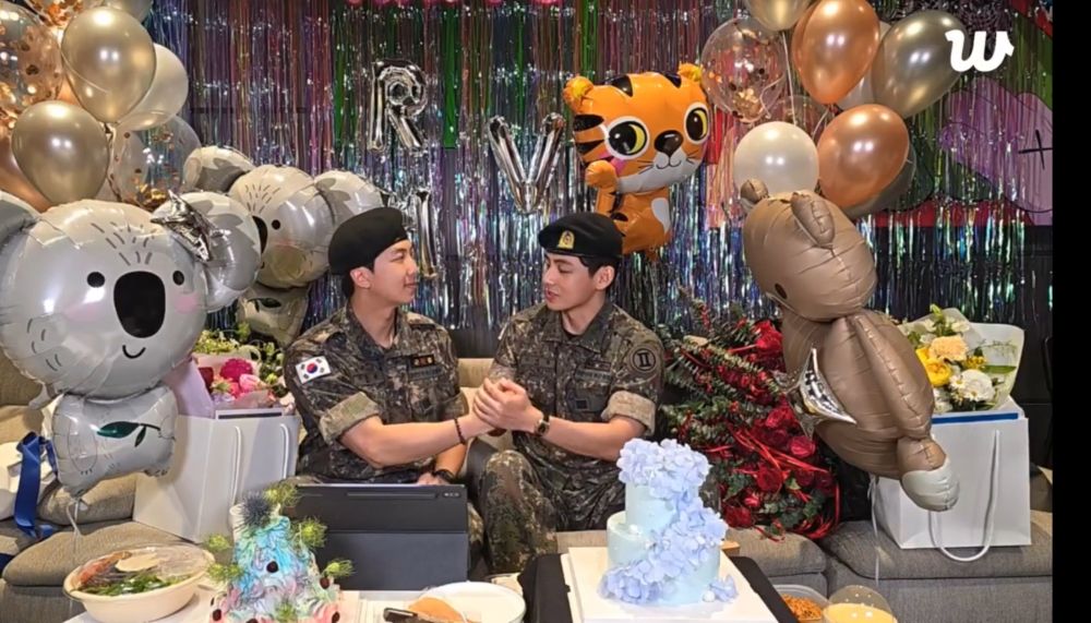 Namjoon and Taehyung sit on a sofa surrounded by festive decorations, gifts, and cakes. They’re in their military fatigues, and have turn toward each other to clasp hands in congratulations for being discharged.