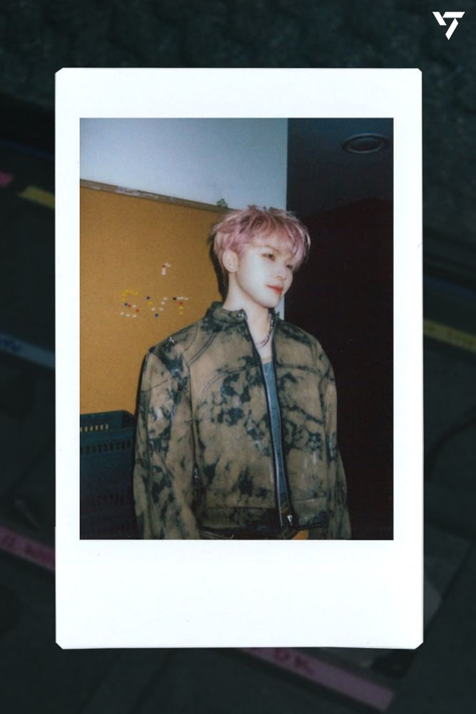 A Polaroid style photo of Woozi of Seventeen in which he has pink hair! An incredible jawline!! And is staring off into the distance unaware of the damage he is causing to my heart. He is standing in front of a cork board on which someone has spelled SVT with pushpins. 