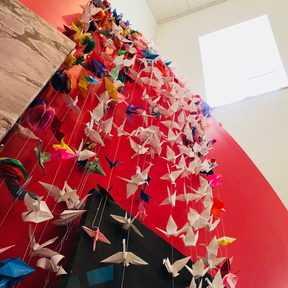 Image looking up at many origami cranes on strings from the ceiling. Bright red wall underneath. Window above with light pouring in. 