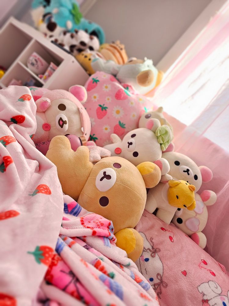 A pile of plushies on a pink, strawberry themed bed. The plushies are primarily of Rilakumma and Korilakkuma. There is a shelf in the background with more collectibles on it as well.