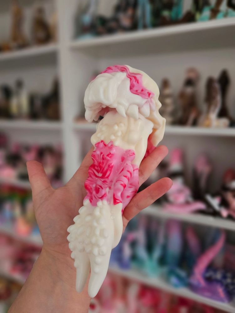 A fantasy sex toy held up to the camera in the palm of the photographer's hand. It is a dragon's mouth with a long, spikey tongue pointing towards the viewer. The dildo was poured in colors resembling a strawberry shortcake. There are more dildos on the shelves behind the toy that is in focus.