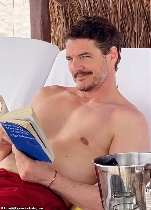 Pedro Pascal shirtless in a red baiting suit reading a blue book at a beach resort.