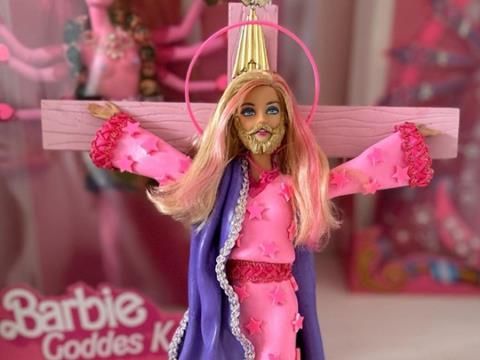 Barbie looking like Jesus Christ with a beard on a pink crucifix wearing a pink rob, and purple cape.