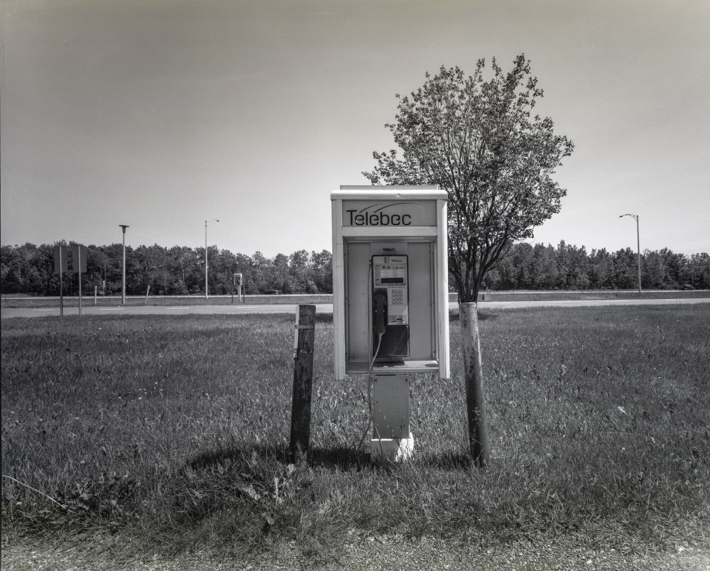 A phone kiosk which is just barely off the ground. Two bent pipes flank it as protection. A tree is immediately behind and an extra tall kiosk for truckers is in the distance.