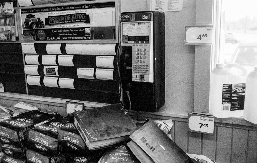 A wall mounted Bell payphone. beneath it are two black binders containing telephone directories that are resting on bags of Kingsford charcoal and Windsor water softener salt. To the left is a bulletin board with small cards for community notices topped by an ad for an auto garage and used car dealer.