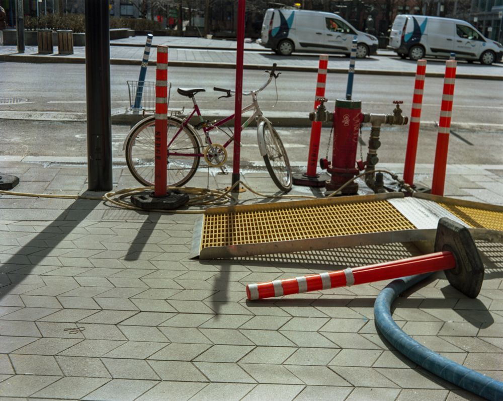 A backlit scene of a sidewalk with zig zagging paving stones. A blue temporary water main hose is snaking across it. A yellow ramp for pedestrians is above the hose and surrounded by tall, orange pylons with reflective bands. A bicycle is locked to a post by the curb. 