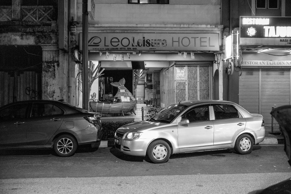 The front of a narrow hotel has a sign reading Leo Leisure Hotel. The font for the word Leisure resembles a cut up Malaysian flag. A man sits at an enormous desk perhaps made from bamboo that resembles a whale. Behind him, a person is looking out from a staircase. A silver car is very poorly parked out front.