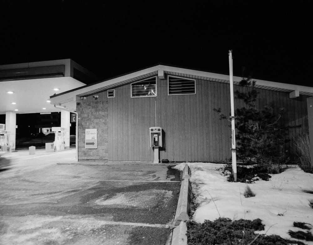 The side of a gas station with a peaked roof and vertical siding. a Bell Canada pay phone kiosk is dead center. In front are parking spots covered with snow, ice and salt stains. Partly melted snow covers most of a small lawn with a tree and perhaps a flagpole. Behind is a large, brightly lit canopy over gas pumps.