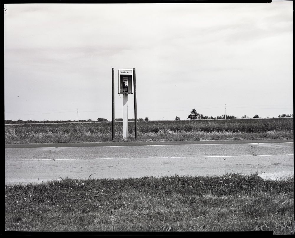 A Télébec payphone kiosk about two metres tall sits next to a rest area road. Steel pipes on either side guard it from motorists. A highway and a line of trees is in the distance.
