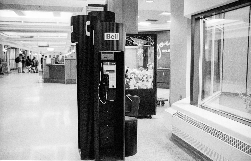 Three cylindrical Bell pay phone kiosks near a large window. Each is a slightly different height. A very long room is behind them, as is a claw machine.