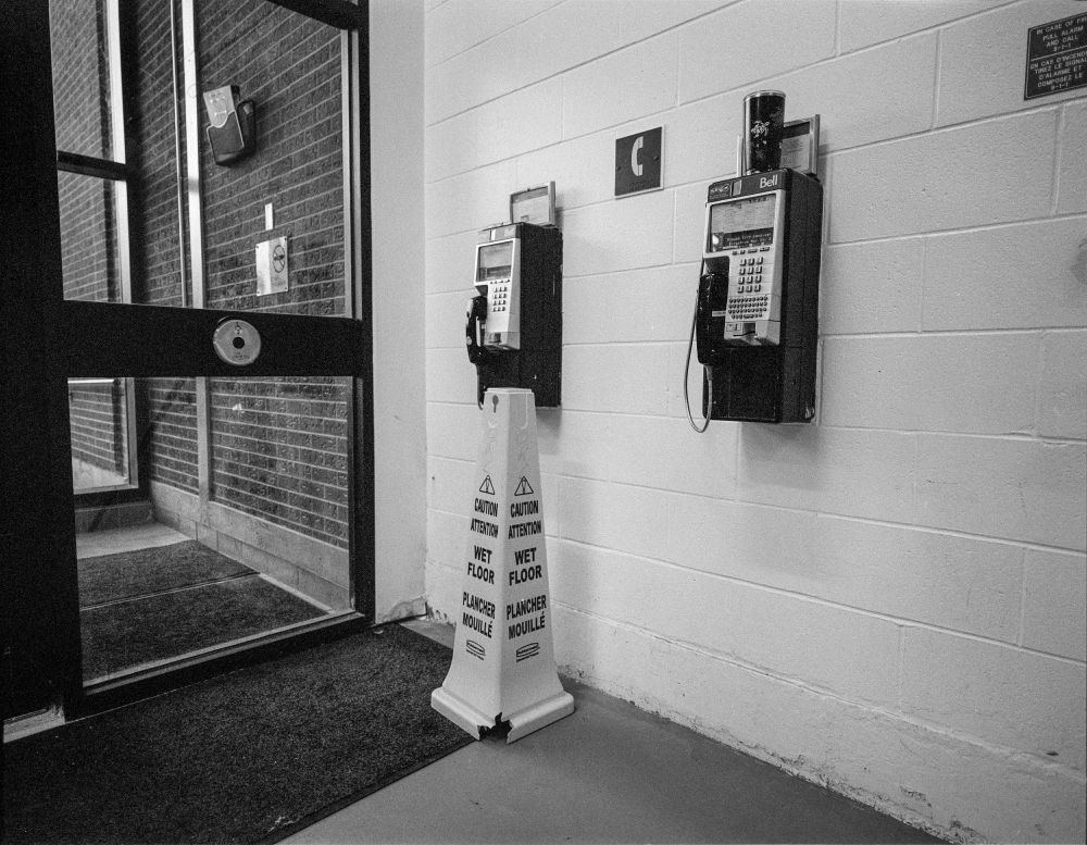 Two payphones mounted on a painted concrete block wall next to a entranceway. In between them is a pictograph sign of depicting a telephone handset. An insulated drinks mug is sitting atop the phone on the right. A tall, three sided pylon warning of a wet floor is in front of the phones. 