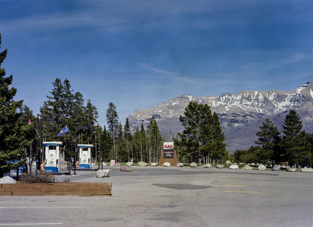 Two large gas pumps both with flags mounted an an angle: one Canadian, one Albertan. A sign says GAS in large red letters and Saskatchewan River Crossing in small type underneath. A circle of large rocks is ahead of the sign, evergreen trees are behind it. Further behind are large mountains with streaks of snow.