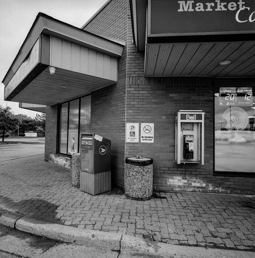 A Bell payphone kiosk near the corner of a store. Instead of a right angle, the corner is a 45 degree wall. There is also two trash bins and a Canada Post mail box. A sign in the shop window gives the estimated prizes for two lotteries: $20 millon and $12 million. A sign on the shop reads: Market with another word that is cut off except for a C.
