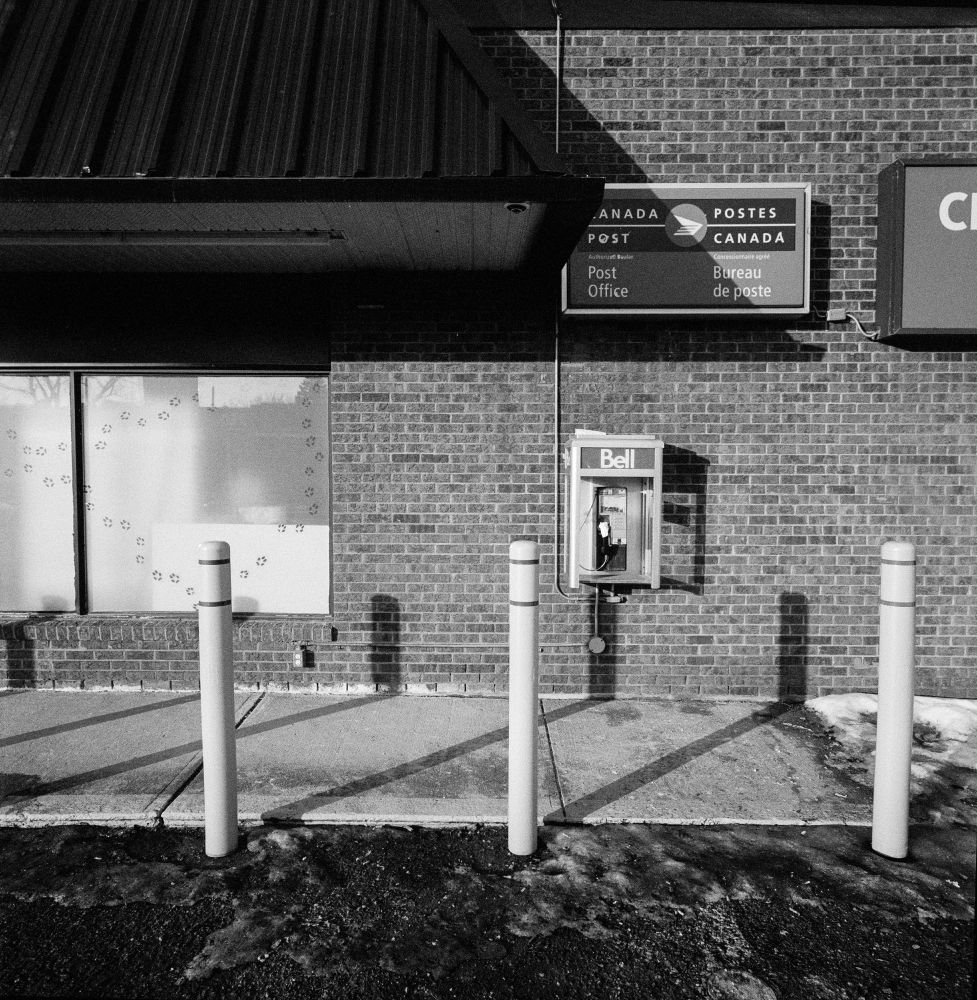 A Bell Canada payphone kiosk mounted on a brick wall under a Canada Post post office sign. Three tall bollards are in front of it and there some melting snow on the ground. To the left are shop windows with some sort of mirrored coating.