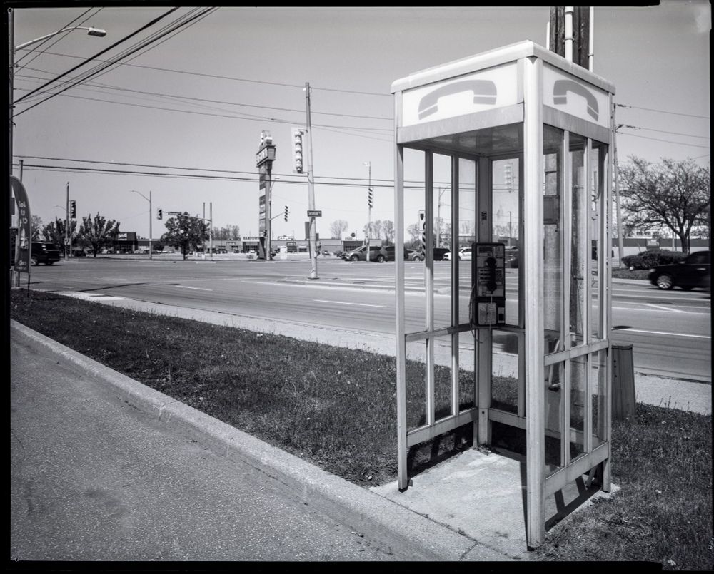 A phone booth in bright sun. Its signs are handset pictographs. Behind is an intersection of two wide roads with a large sign looming and a shopping mall in the distance. 