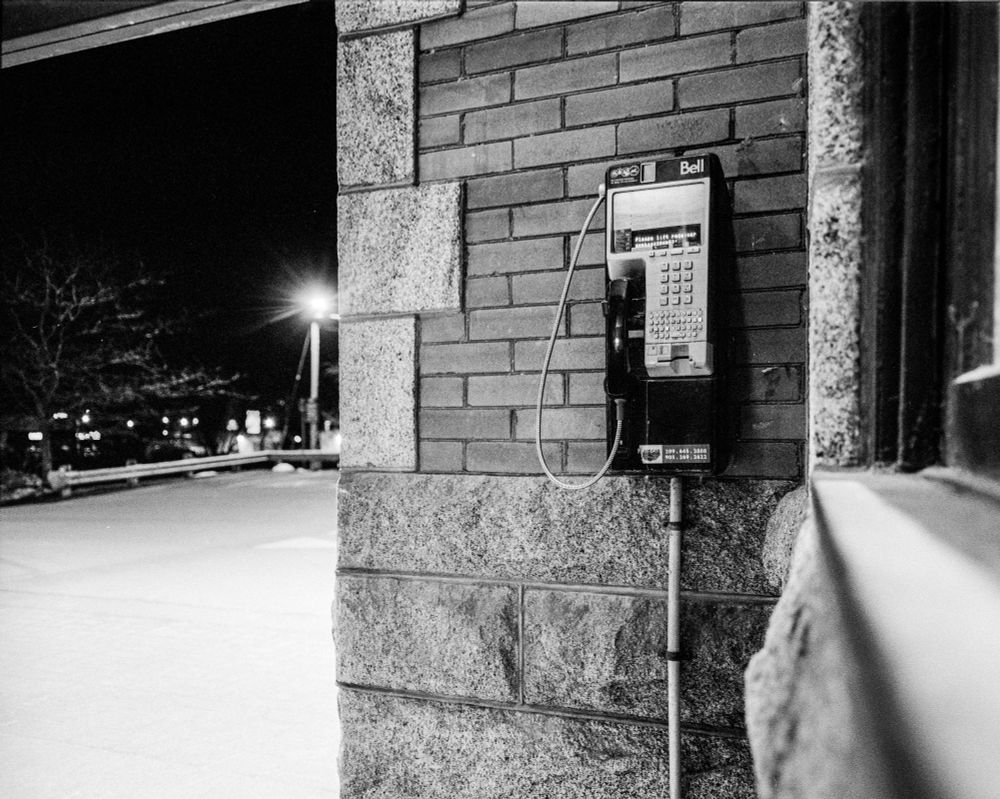 A Bell payphone mounted on a brick wall which has carved stone elements. It is at night and a lamp is illuminating an empty part of a parking lot.
