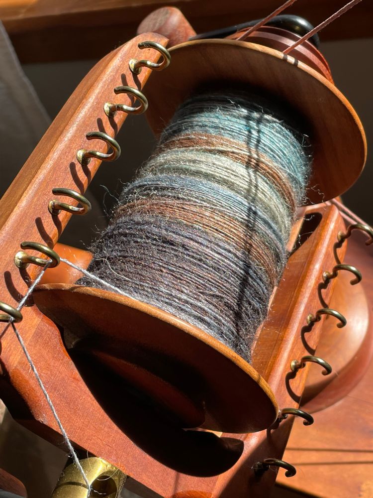 A cherry Lendrum saxony wheel bobbin and flyer fill the frame with intense sunshine and shadows from the driveband visible. There’s yarn in shades of gray, purple, copper, black, blue and pale green visible as singles on the bobbin which is mostly full. 

#handspinning #yarn 