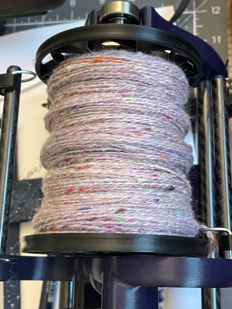A dark blue electric spinning wheel is in the foreground with a mostly-full black bobbin of light cool pink yarn spun and plied. In the background is paper and an inch-grid cutting mat.

#handspinning #handspun #sarisilk #knitsky #spinning #spinnersofbluesky #wool #yarn #daedalusspinningwheels #daedalussparrow 