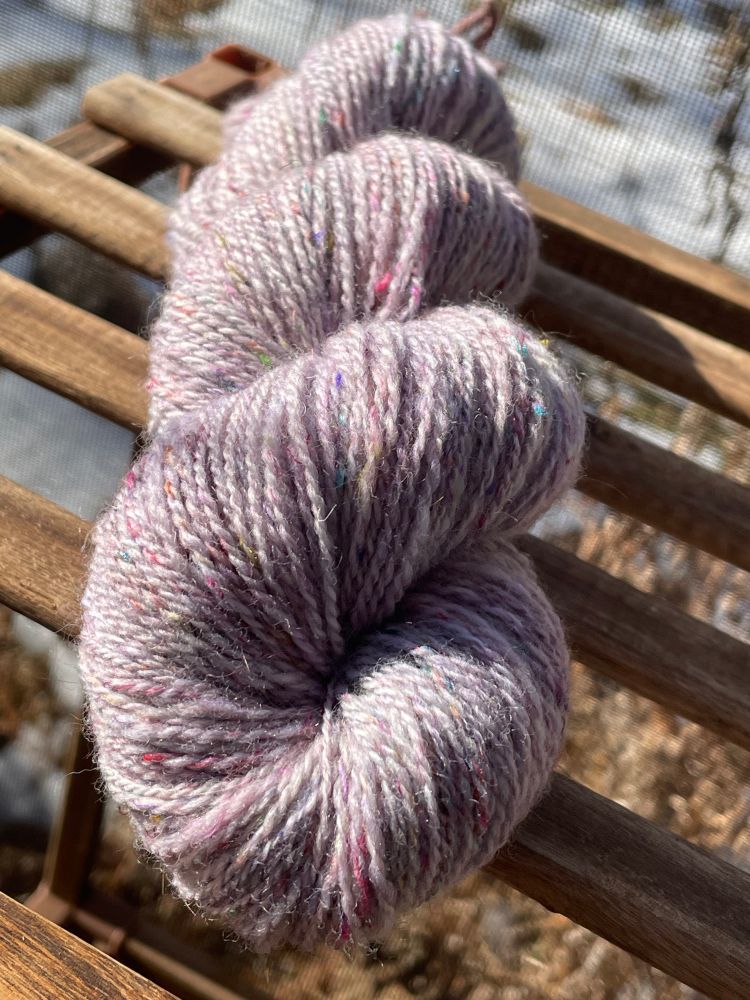 A twisted skein of handspun yarn sits on a wooden frame with natural light creating high contrast lights and shadows. The color of the yarn is a pale cool pink with bits of colorful silk threads, approximately sock or fingering weight with two plies. The background is full of leaves and snow, with a screen texture visible in the background. 

#handspinning #handspun #sarisilk #knitsky #spinning #spinnersofbluesky #wool #yarn #fiberarts #fibrearts