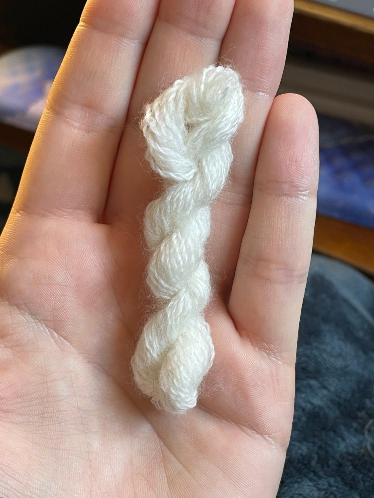 A small several inch long twisted skein of glowy white wool lays in the hand of the photographer. A desk and keyboard are seen out of focus in the background. 