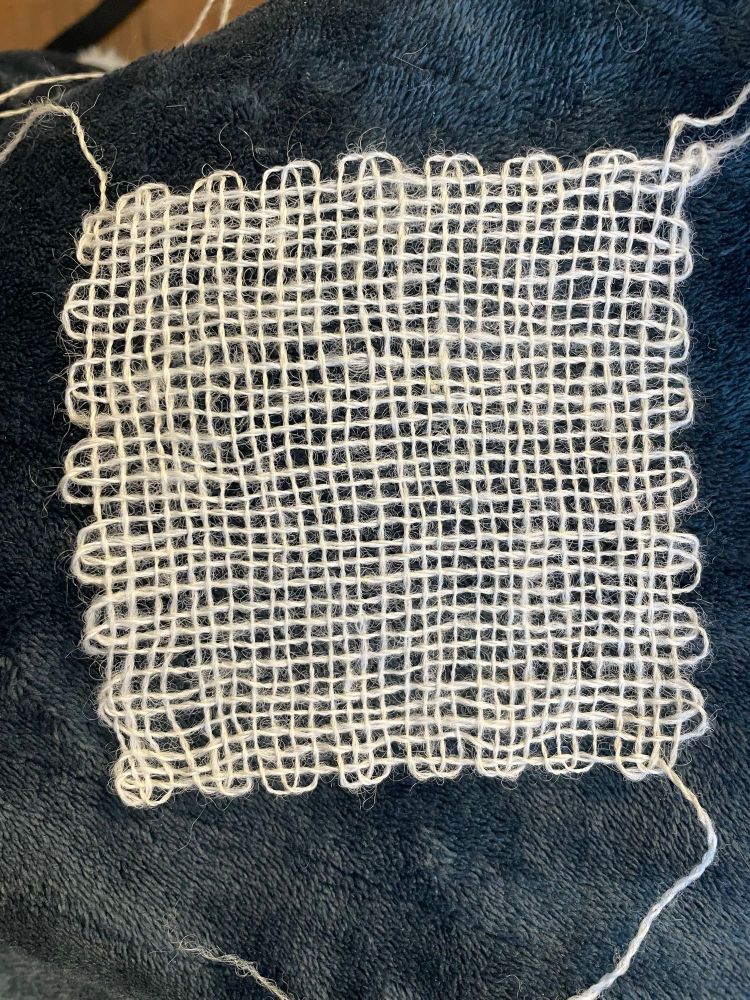 A sample square of the fine two ply Shetland yarn woven on a handheld 4” loom sits on a blue fuzzy blanket. Multiple loose ends of wool are visible. The texture of the square is loose and lacey, highlighting the twist and texture of the yarn.

#shetlandwool #rawfleece #spinning #wool #yarn #weaving 