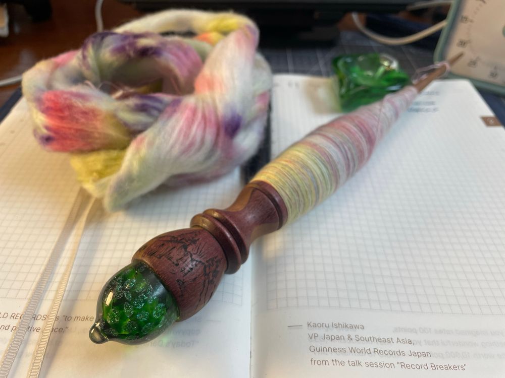 A Hobonichi planner sits open to Feb 11th. A green glass tipped richly colored and woodburned turned spindle sits on the open planner with a cop of finely spun pastel colored cotton visible but out of focus. A wound nest of white cotton with splotches of pink, purple and yellow is visible to the left and a green glass heart (used as a bowl for spinning) is visible to the right. A blue timer is also just out of frame to the top right.
#handspinning #yarn #spinning #supportspindle #cottonspinning #mingoandasho 
