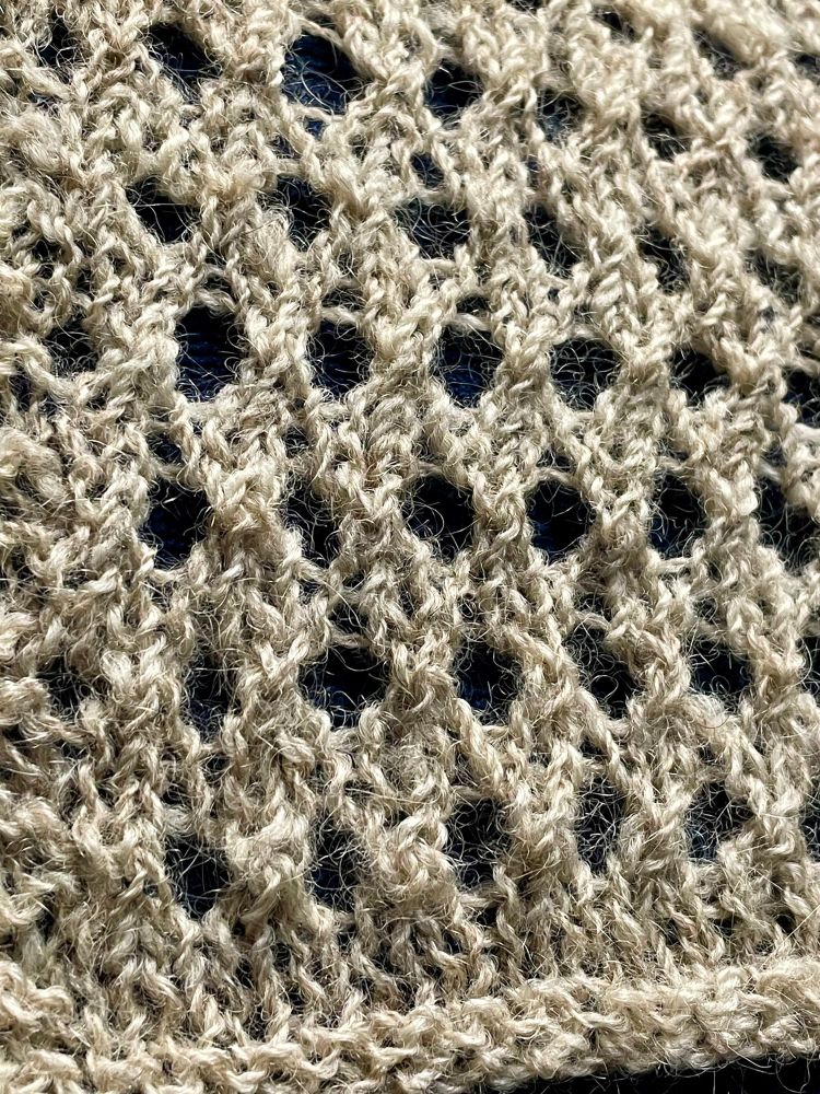 A closeup picture of a knitted lace swatch with a shiny beige yarn with halo visible in the lace pattern yarn-overs. 
