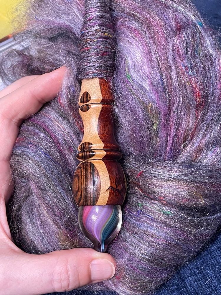 A batt of sparkly purple fiber with glossy silk bits and threads sits on a blue pillow with a hand holding a wooden spindle made of several distinct types of wood turned in a decorative pattern that’s topped with a glass tip swirled in purple, pink and green on top. A small cop of 🧶 yarn is visible wound on the shaft of the spindle.

Spindle by #mingoandasho and fiber blended by an artist not yet on BlueSky. 

#Spindlespinning #supportspindle #fiberarts #spinning 