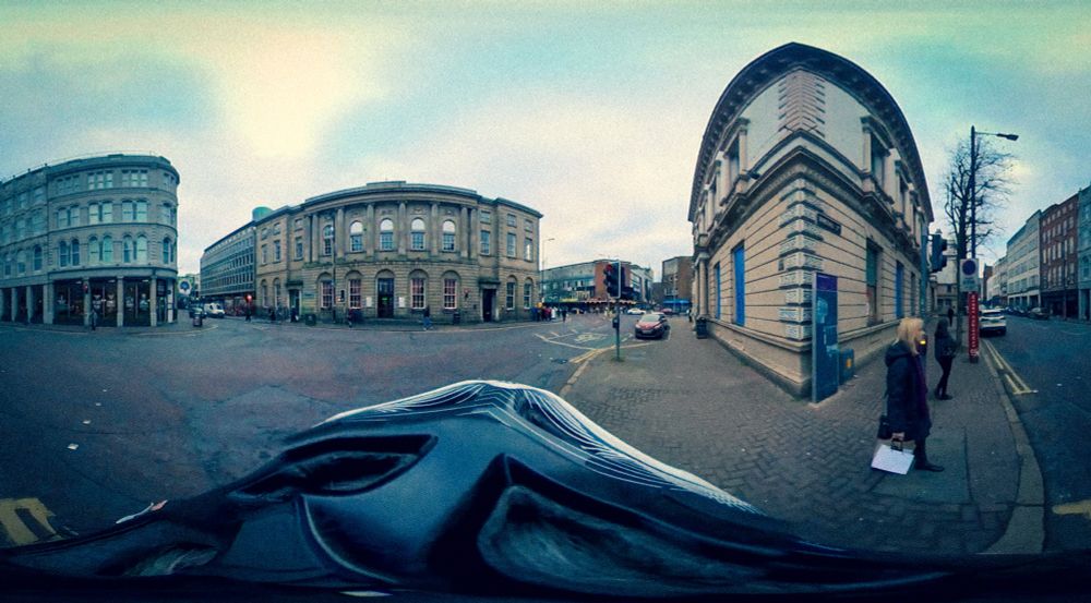 Equitectangular stretched image showing buildings in Belfast city centre, some pedestrians and a bicycle helmet occupying the lower section of the image. 