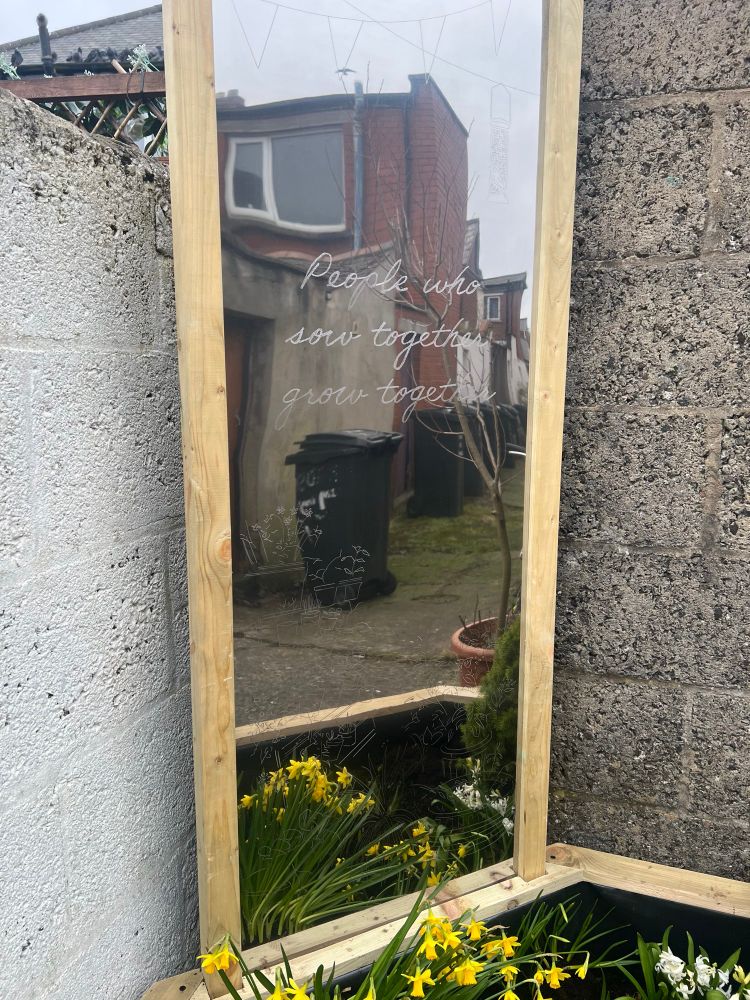 A closer view of the upper section of the mirror, featuring a delicate etched design of bunting and architectural details. The reflection captures the alleyway behind, showing bins, a bicycle, and red-brick buildings.
