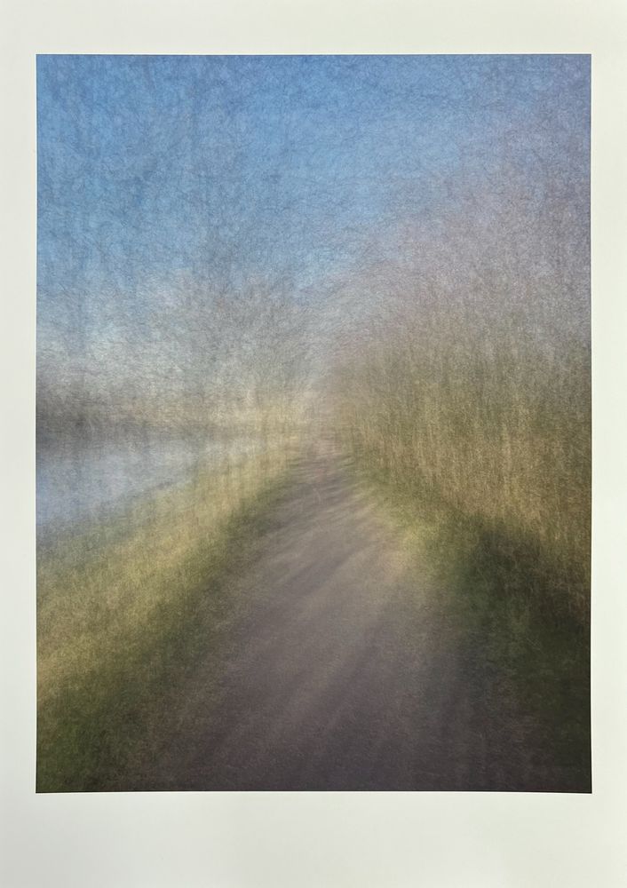 An abstract, blurred image of a winding path alongside a body of water under a soft blue sky. The path, light gray with some darker streaks, curves gently from the lower center towards the upper right. To the left of the path, a calm body of water, depicted in muted blues and grays, reflects the sky. To the right, a line of trees or vegetation, blurred with horizontal streaks of light brown and green, forms a soft barrier. The sky above is a broad expanse of soft blue, with a hazy, textured appearance. The image conveys a sense of peaceful movement through a natural landscape.