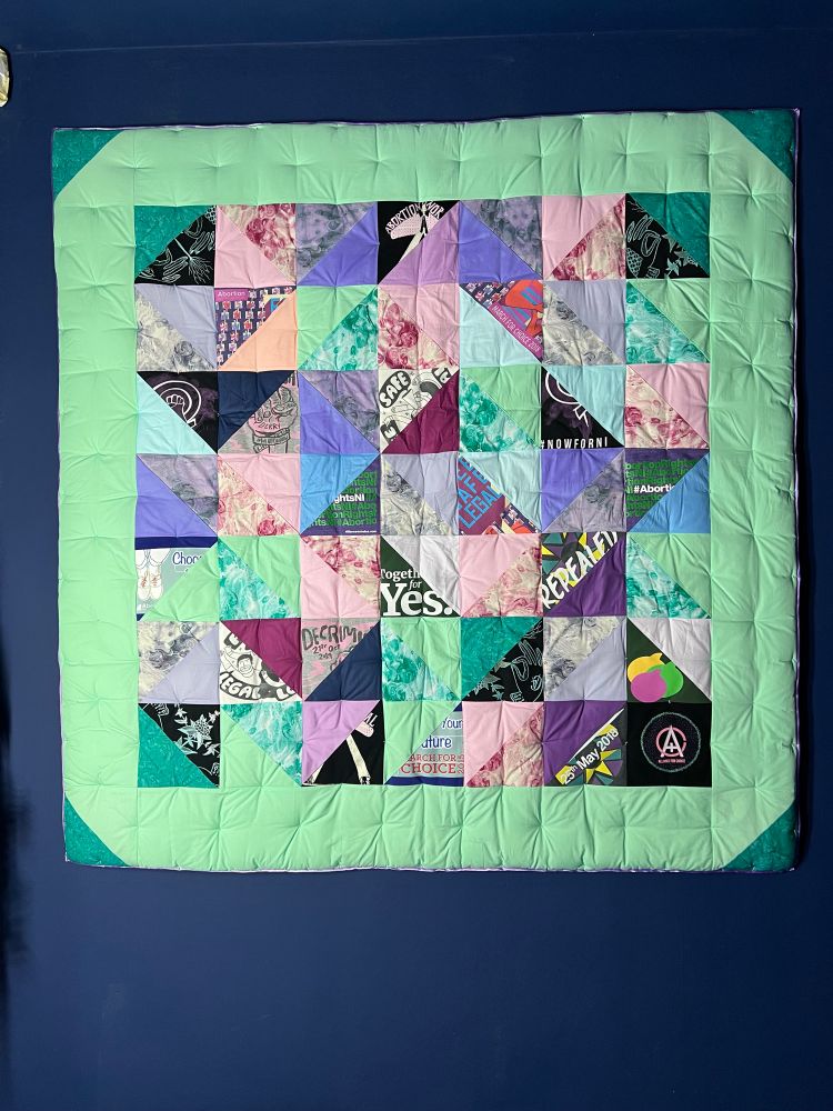 Quilt on a Dark Blue Wall – A large, square quilt with a geometric patchwork design is displayed against a dark blue wall. The quilt features a mix of pastel and vibrant fabric pieces, including sections with text and graphic designs. The border is a soft green color, framing the central pattern of triangles and squares.