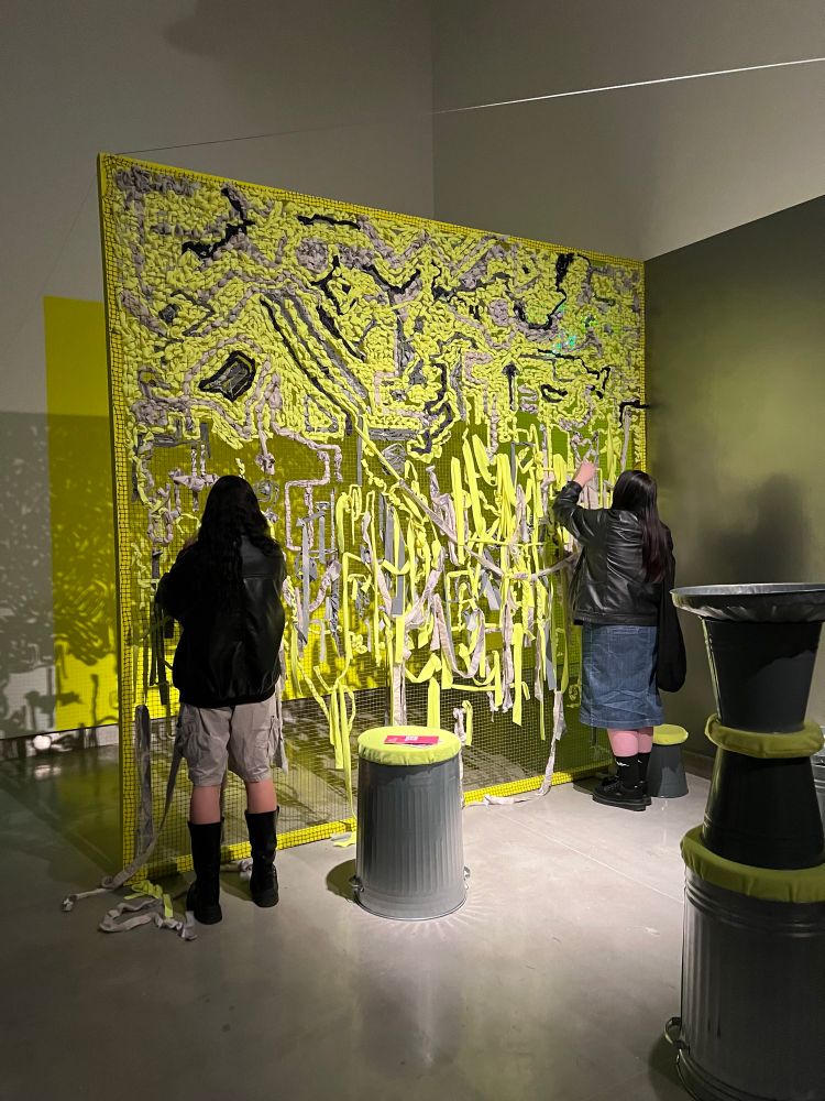 Two people embroider using pieces of neon and grey fabric a large net. Surrounding them are various bins with yellow neon covers. 