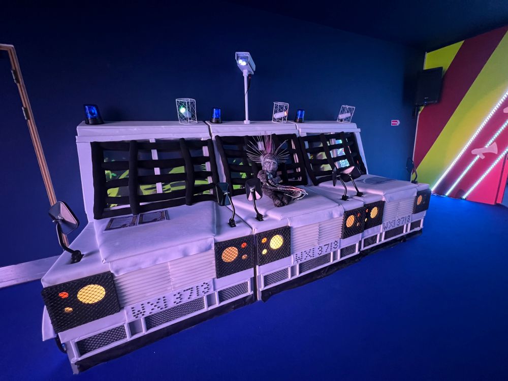 Police Van-Inspired Seating Installation – A seating installation designed to resemble a row of armored police vans is displayed in a dimly lit room with dark blue walls and floors. The van-like seats are white with black grilles and metal details, and each has a blue flashing light on top. A sculpted figure with a punk-inspired headdress sits on one of the seats.