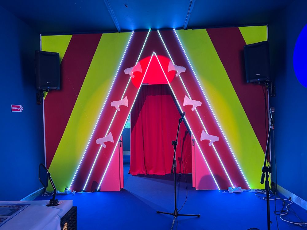 Stage Entrance with Megaphone Motif – A theatrical stage entrance is designed with bold diagonal stripes in red, yellow, and green. White megaphone-shaped cutouts are mounted along the entrance, accompanied by LED strip lights. Two microphones stand in front of the entrance, which is draped with red fabric.