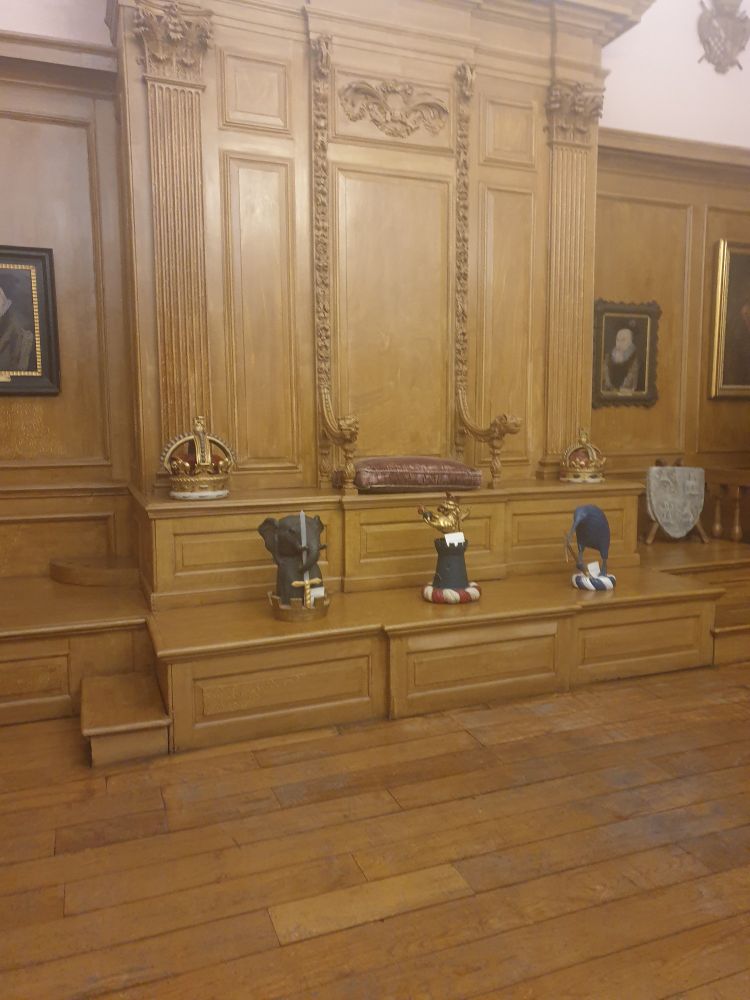3 ornamental Caps of Maintenance on a wooden dais,  with 2 crowns and cushioned seat above, with wooden panelling on wall and floor 