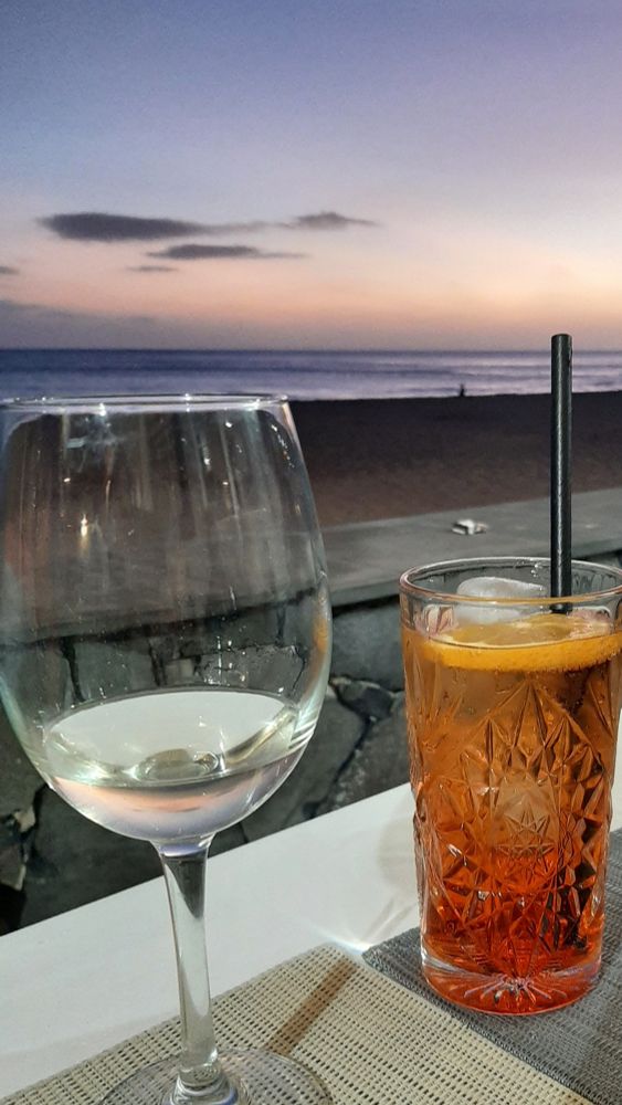Strandszene, im Hintergrund dss Meer kurz nach Sonnenuntergang, im Vordergrund ein weißer Tisch an einer Strandpromenade, ein Glas mit Weißwein ein Longdrinkglas mit rötlicher Flüssigkeit, Eiswürfeln, Orangenscheibe und Strohhalm.
