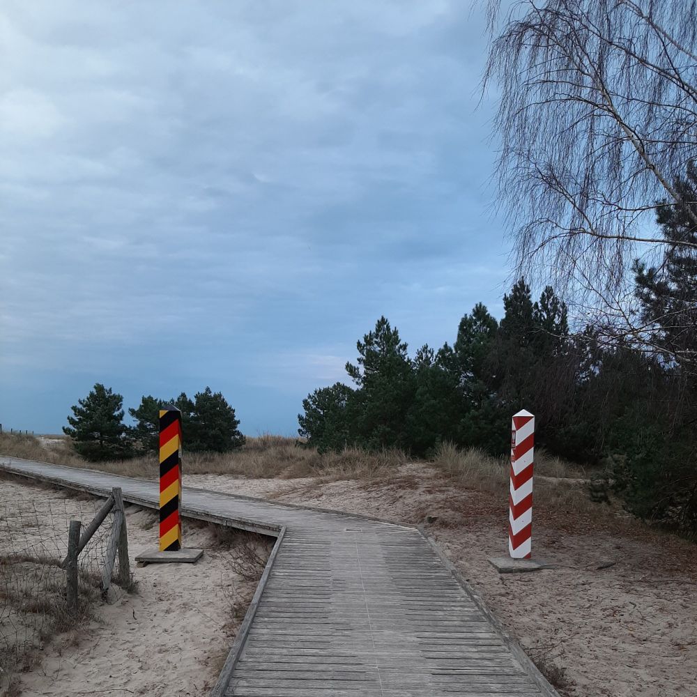 Grenzübergang Polen-Deutschland in Ostseenähe. Ein langer Holzsteg durch eine Dünenlandschaft, links ein  schwarz-rot-goldener Pfosten, rechts einer in rot-weiß.