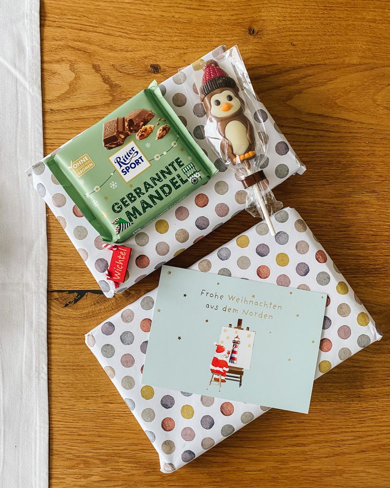 Zwei verpackte Bücher, eine Postkarte, eine Tafel Schokolade und ein Schokololli liegen auf einem Tisch.