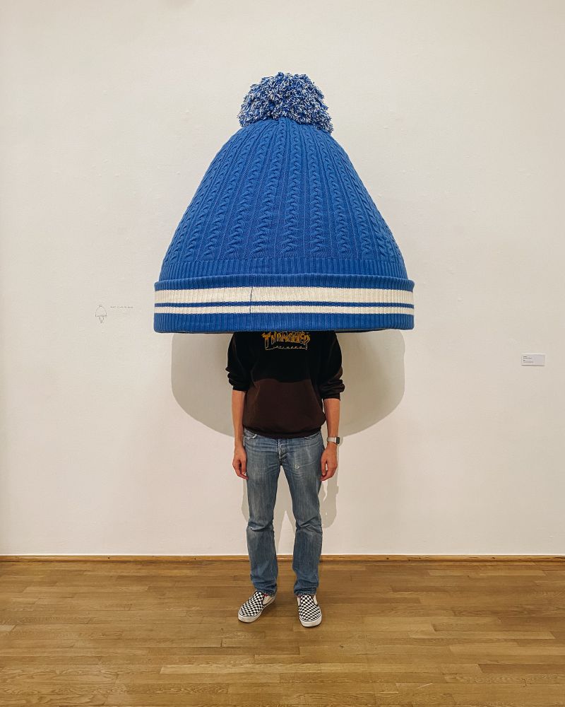 Ein Mensch steht in einem Museum unter einer überdimensionalen Strickmütze.