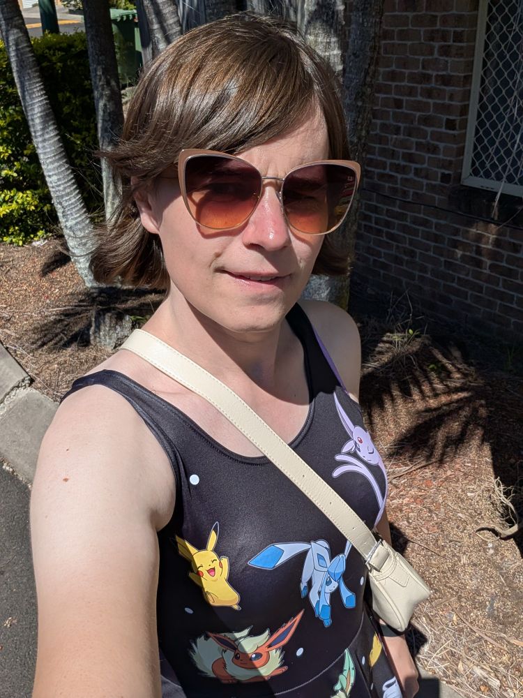 Selfie of me outside in bright sunshine, wearing a black dress with various Pokemon on it.