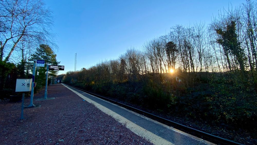 A photo taken at Connel Ferry railway station. The rising sun is peeking through spindly autumn trees. An electronic notice board reads “Glasgow Queen Street”.
