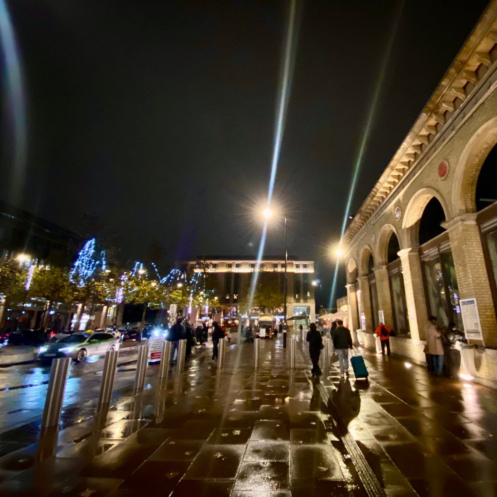 Snapshot of the concourse in front of Cambridge railway station. It’s dark and the pavement is slick with rain. 