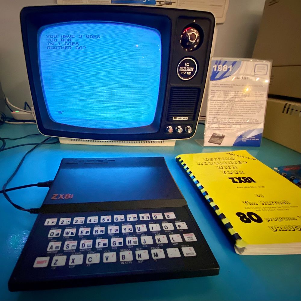 Photograph of a museum display featuring a ZX81 computer and a small portable black-and-white television. Text on screen reads: “you have three goes. You won in 1 goes. Another go?”