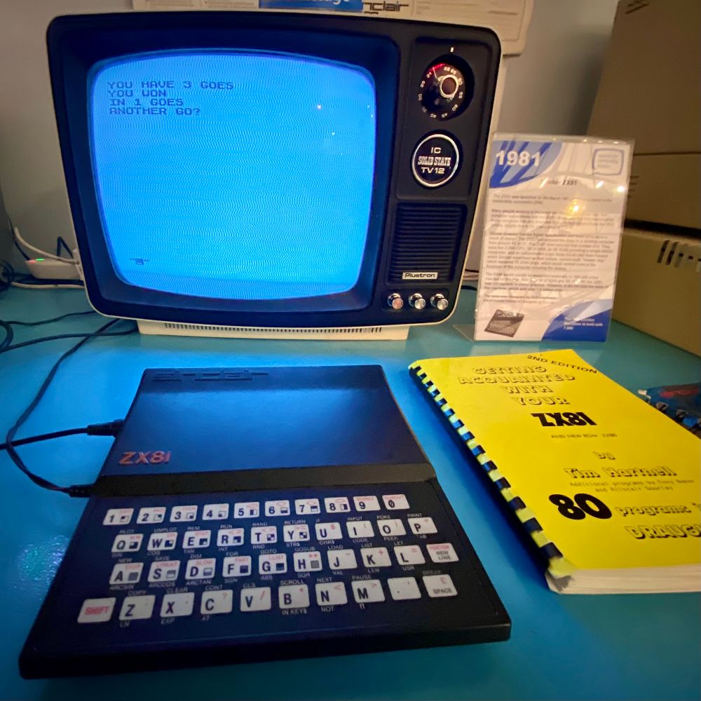 Photo of a museum exhibit. A ZX81 and a portable black and white telly. Text on the TV reads “You have three goes. You won. In one goes. Another go?”